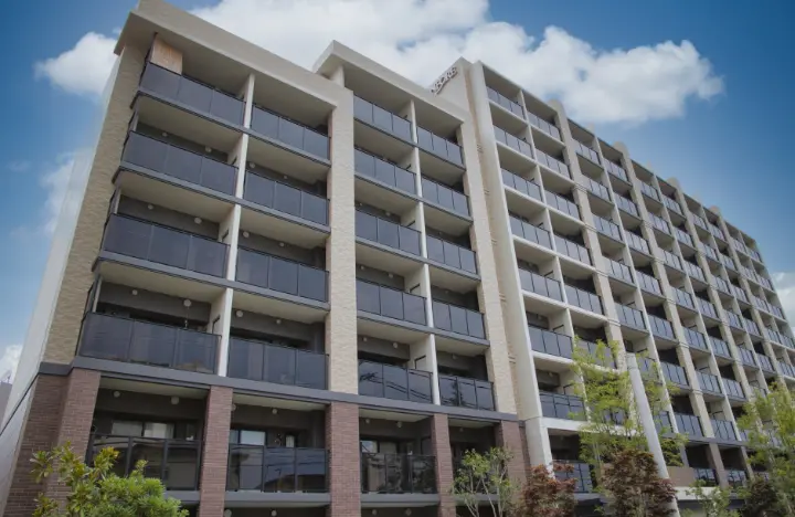 緑の植栽が並ぶエントランス付近から見上げたマンション全景の写真